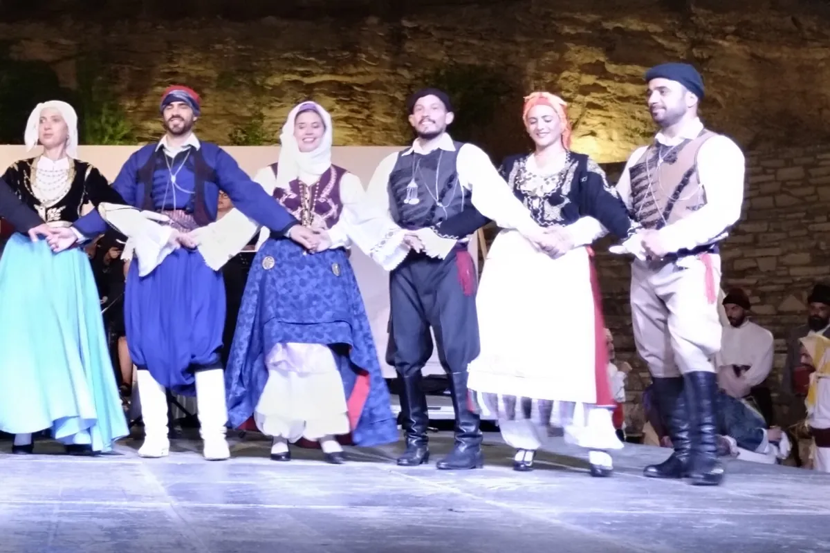Dancers in Cretan costume with linked hands: men in dark baggy breeches and boots, and women in long skirts, embroidered bodices and white headscarves