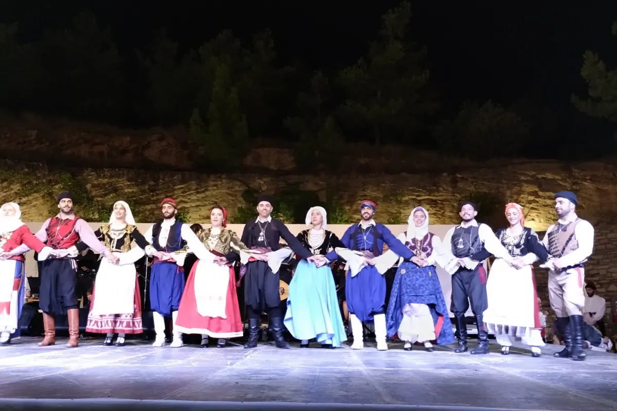Dancers in Cretan costume standing in a line with linked hands on an open-air stage at night, in front of a stone terrace and pine trees