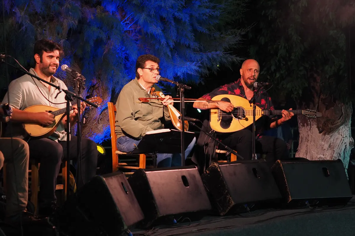 Three musicians on an open-air stage at night playing a mandolin, a Cretan lyra and a laouto, under blue-lit trees